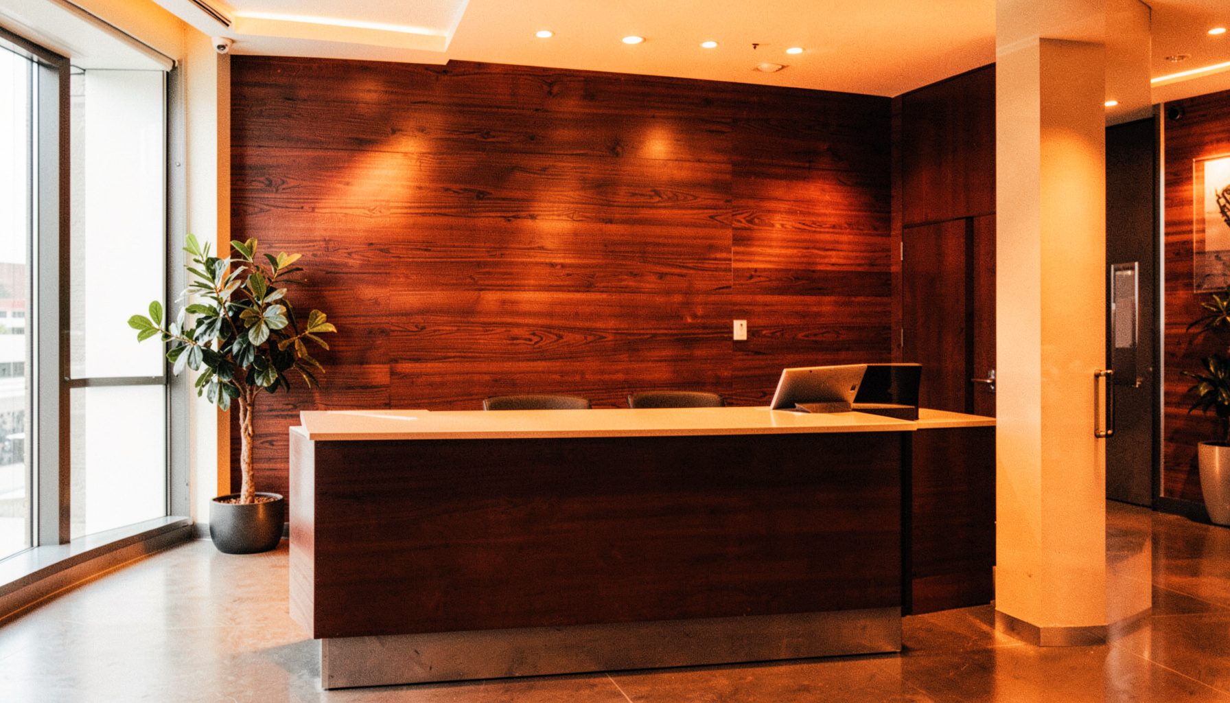 Warmly lit corporate reception area with a wood-textured accent wall in rich brown tones, a sleek desk, and soft ambient lighting conveying a professional and welcoming atmosphere.